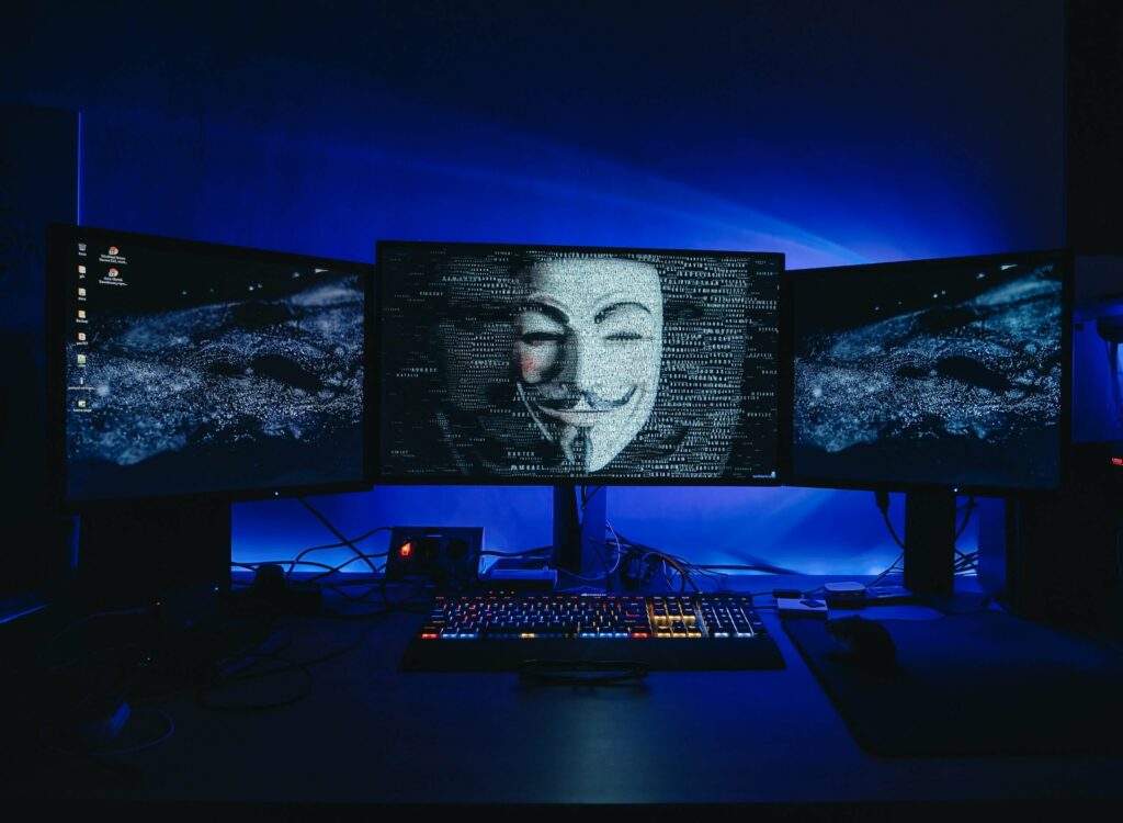 Three computer screens with anonymous mask on center screen.